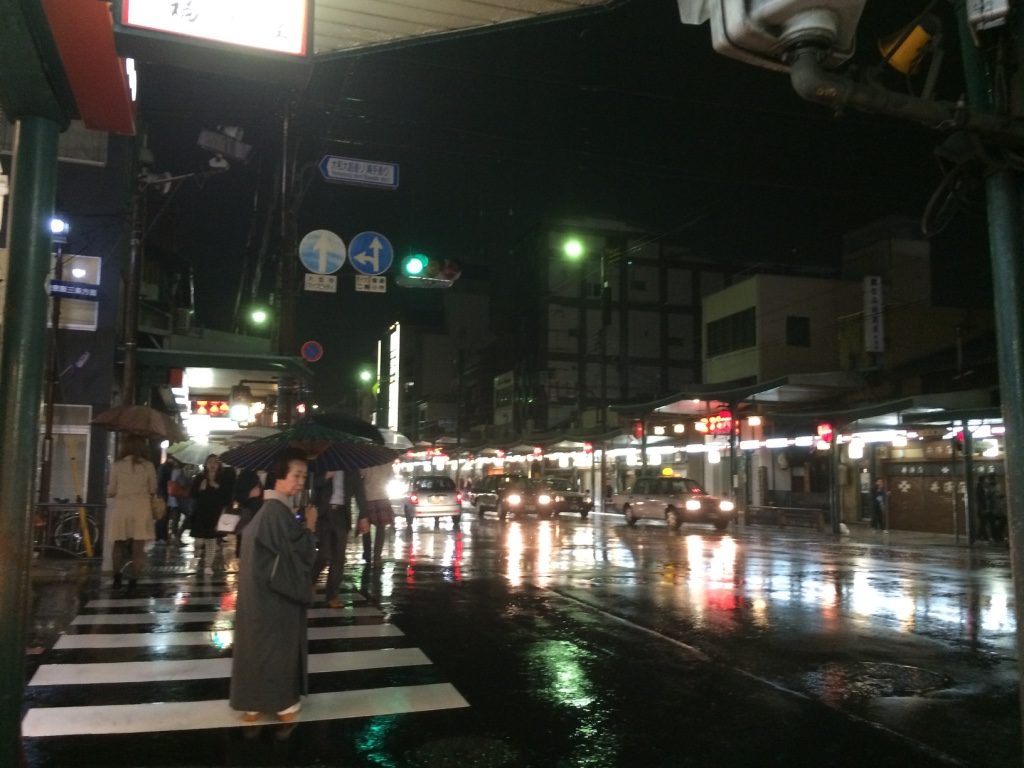 Older lady in traditional Japanese attire and umbrella trying to get a taxi. After waiting for a little bit, a young Japanese man goes up to her and asks her if she needs help and he within a couple minutes he manages to wave a cab down for her. 